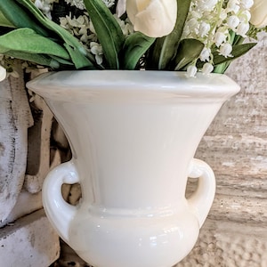 Puede incluir: Un jarrón de cerámica blanco con dos asas, lleno de flores blancas y hojas verdes. El jarrón tiene una forma de urna clásica y se apoya sobre una superficie blanca. Las flores incluyen tulipanes y otras flores blancas.