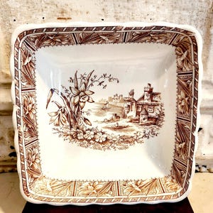May include: A square, white ceramic dish with a brown transferware design. The center features a landscape with a building and flowers. The rim has a decorative floral pattern. The dish has a scalloped edge.