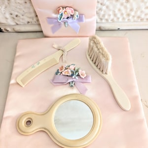 May include: A vintage vanity set in soft pink, featuring a comb, brush, hand mirror, and a small pouch. Each item is cream-colored and adorned with floral accents and ribbons. The set is arranged on a pink satin surface.
