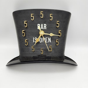 May include: Black novelty clock shaped like a top hat, with gold-colored hands and numerals. The clock face reads "BAR IS OPEN" and features the number 5 repeated around the perimeter. The clock is a vintage item.