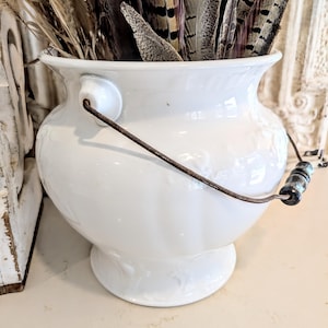 May include: A white ceramic pot with a rounded body and a flared rim, featuring a rusted metal swing handle with a dark wooden grip. The pot is filled with a collection of brown and black patterned feathers and dried grasses.