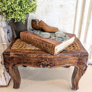 May include: A small, antique wooden table with carved legs and a woven top. A vintage book, reading glasses, and a wooden shoe form rest on the table. A potted plant sits to the left, creating a vintage aesthetic.
