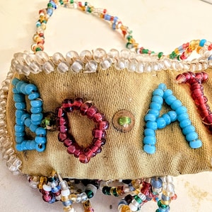 May include: A vintage beaded bag with the word "BOAT" spelled out in colorful beads. The letters are blue and red, set against a tan fabric background. The bag has a beaded handle and trim.