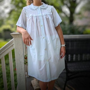 May include: A light blue knee-length house dress featuring a Peter Pan collar with white lace trim, short sleeves, and a front button-down closure. The garment includes a gathered yoke, two side pockets, and delicate white floral embroidery.