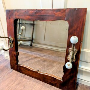 May include: A rectangular wall mirror with a dark brown stained wooden frame. The mirror features two brass-toned metal coat hooks attached to the sides, each equipped with two white spherical knobs at the ends of the curved metal arms.