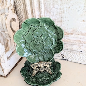 Può includere: Due piatti verdi in ceramica a forma di foglia. Il piatto superiore è più grande e presenta un motivo a foglie dettagliato. Il piatto inferiore è più piccolo e ha un elemento decorativo in metallo color argento. I piatti sono su una superficie chiara.