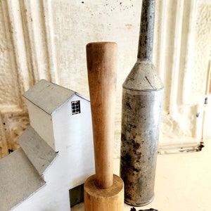 May include: A collection of vintage farmhouse decor. Includes a wooden butter churn, a metal funnel, a miniature white barn, and two small cow figurines. The items are arranged on a cream-colored surface against a textured white background.