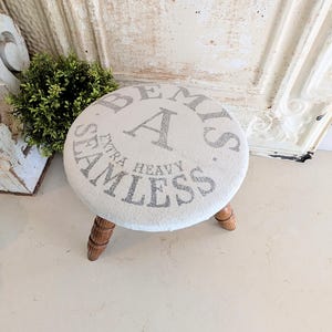 Puede incluir: Un pequeño taburete redondo con asiento crema y texto gris "Bemis Extra Heavy Seamless". Tiene tres patas de madera torneada. El diseño es de estilo vintage.