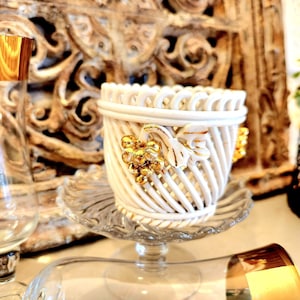 Puede incluir: Un recipiente blanco de estilo cesta tejida con detalles dorados, con un diseño floral dorado. El recipiente se encuentra sobre una fuente de pedestal de vidrio transparente. Decoración del hogar.