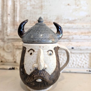May include: A ceramic mug featuring a sculpted face with a prominent nose, wide eyes, and a dark brown mustache. The mug includes a removable lid shaped like a grey, speckled Viking helmet with two curved horns extending from the sides.