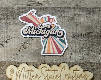 May include: A sticker featuring a retro-style illustration of the state of Michigan with the word "Michigan" in a stylized font. The sticker is on a wooden surface with a wooden sign that reads "@Mitten State Crafting".