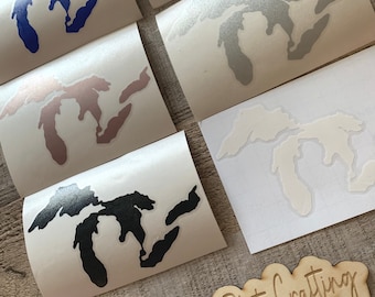 May include: A set of six Great Lakes outline decals in various colors, including black, white, silver, pink, brown, and blue. The decals are on white paper and are ready to be applied to a surface. The decals are displayed on a wooden surface with a sign that reads "@Mitten State Crafting".