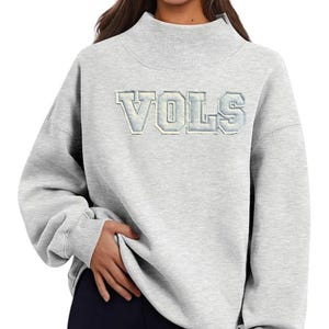 Puede incluir: Sudadera gris claro con cuello alto y mangas largas. La palabra "VOLS" está bordada en el pecho en azul claro y blanco. La sudadera está hecha de un material suave.