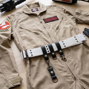 May include: A tan jumpsuit featuring a Ghostbusters logo patch on the right sleeve and a black name patch with the text VENKMAN on the chest. A white webbed belt with black leather straps, metal snaps, and hanging metal rings is worn over the jumpsuit.
