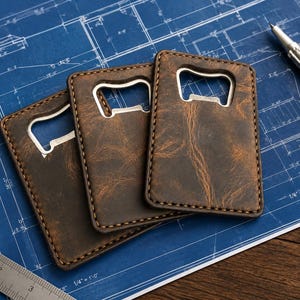 May include: Three brown leather bottle openers with silver metal openers are arranged on a blue blueprint. The leather has a distressed texture and is stitched around the edges. A silver pen and a metal ruler are also on the blueprint.