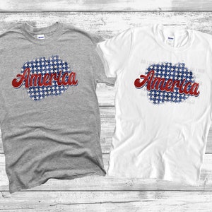 May include: Two short-sleeved t-shirts, one gray and one white, are displayed. Each shirt features the word "America" in red script over a blue and white starburst design. The shirts are likely made of cotton.