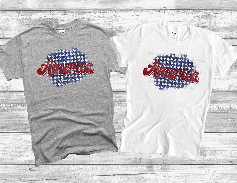 May include: Two short-sleeved t-shirts, one gray and one white, are displayed. Each shirt features the word "America" in red script over a blue and white starburst design. The shirts are likely made of cotton.