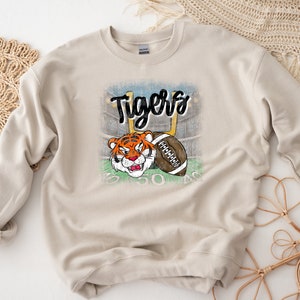 May include: A beige sweatshirt with a distressed graphic of a tiger holding a football. The graphic includes the word "Tigers" in black lettering.