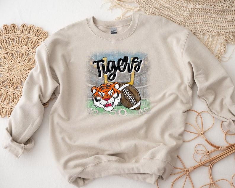 May include: A beige sweatshirt with a distressed graphic of a tiger holding a football. The graphic includes the word "Tigers" in black lettering.