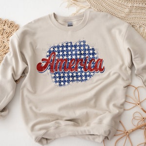 May include: A beige sweatshirt with the word "America" in red and blue script. The word is superimposed over a blue and white star pattern, evoking a patriotic theme. The sweatshirt is a casual garment.