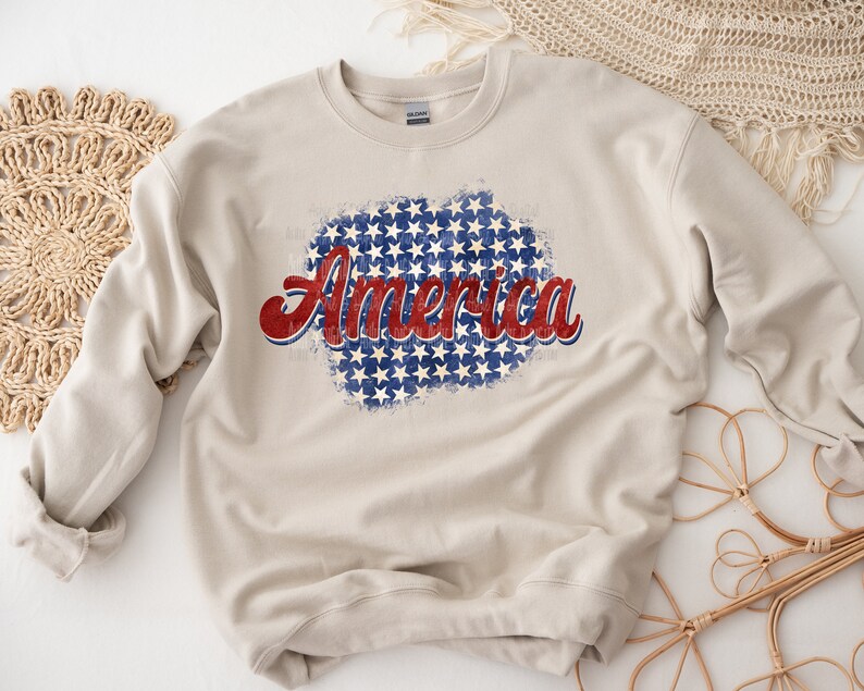 May include: A beige sweatshirt with the word "America" in red and blue script. The word is superimposed over a blue and white star pattern, evoking a patriotic theme. The sweatshirt is a casual garment.