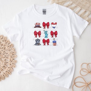 May include: White t-shirt with a red, white, and blue patriotic design. The design includes a liberty bell, a Statue of Liberty, fireworks, a heart with the American flag, and a bow.