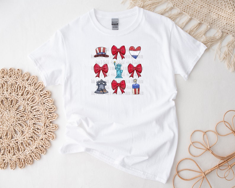 May include: White t-shirt with a red, white, and blue patriotic design. The design includes a liberty bell, a Statue of Liberty, fireworks, a heart with the American flag, and a bow.
