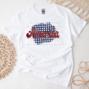 May include: White t-shirt with the word "America" in red and blue script, set against a blue background with white stars. The shirt is a classic crew neck style.
