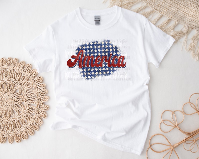 May include: White t-shirt with the word "America" in red and blue script, set against a blue background with white stars. The shirt is a classic crew neck style.