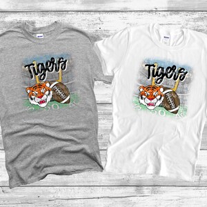 May include: Two t-shirts with a graphic of a cartoon tiger wearing a football helmet and holding a football. The text "Tigers" is above the tiger. The t-shirts are gray and white.