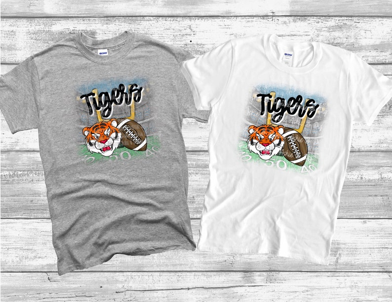 May include: Two t-shirts with a graphic of a cartoon tiger wearing a football helmet and holding a football. The text "Tigers" is above the tiger. The t-shirts are gray and white.