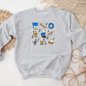 May include: A light gray sweatshirt featuring a collection of cartoon band instruments and musical notes in blue, yellow, and black. The design includes a tuba, trumpet, saxophone, drum, and a marching band leader.
