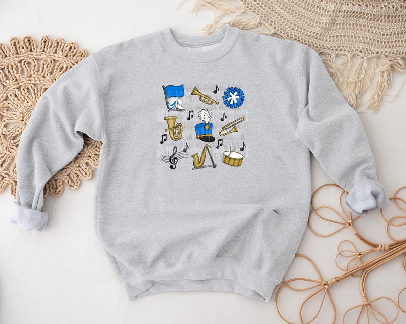 May include: A light gray sweatshirt featuring a collection of cartoon band instruments and musical notes in blue, yellow, and black. The design includes a tuba, trumpet, saxophone, drum, and a marching band leader.