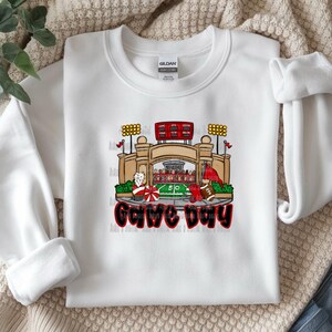 May include: White sweatshirt with a colorful graphic of a football stadium, football players, and the words "Game Day" in red and black. The sweatshirt is made of a soft material.