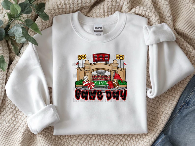 May include: White sweatshirt with a colorful graphic of a football stadium, football players, and the words "Game Day" in red and black. The sweatshirt is made of a soft material.