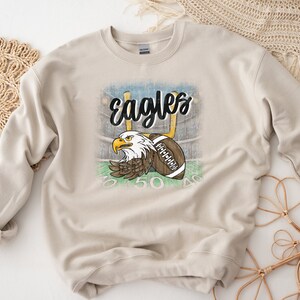 May include: A beige sweatshirt featuring a distressed graphic of an eagle with a football in its talons. The graphic includes the text "Eagles" and football field markings.