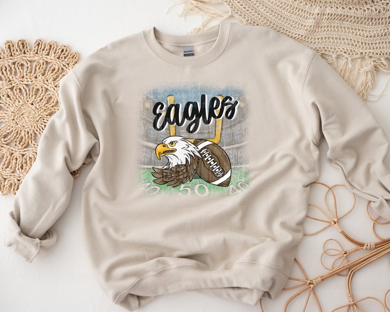May include: A beige sweatshirt featuring a distressed graphic of an eagle with a football in its talons. The graphic includes the text "Eagles" and football field markings.