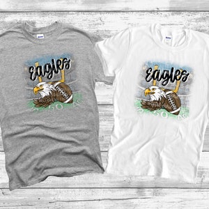 May include: Two t-shirts featuring a graphic design of an eagle holding a football in front of a football pitch. The text "Eagles" is written in a stylised font above the eagle. The t-shirts are grey and white.