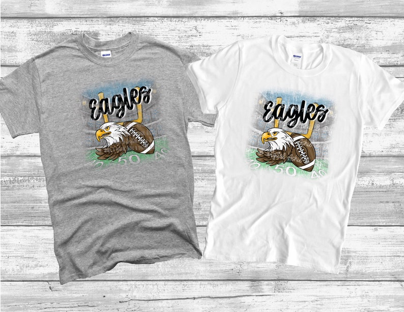 May include: Two t-shirts featuring a graphic design of an eagle holding a football in front of a football pitch. The text "Eagles" is written in a stylised font above the eagle. The t-shirts are grey and white.