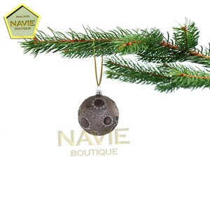 May include: A grey glass Christmas bauble with a moon-like design. The bauble is hanging from a gold string on a green pine branch.