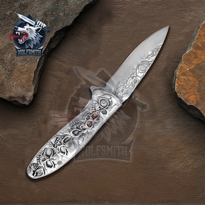 May include: A silver folding knife with intricate skull engravings on the handle and blade. The knife features a pointed blade and a detailed design. The Wolfsmith logo is visible in the upper left corner and on the handle.