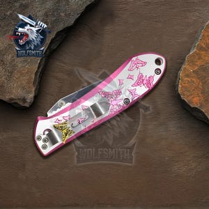 Pu&ograve; includere: Un coltello pieghevole con manico bianco e rosa, decorato con motivi a farfalla rosa. La lama &egrave; argentata e il manico presenta un'emblema dorato. Il coltello ha una clip da tasca e un perno per il pollice. Il logo del marchio "WOLFSMIITH" &egrave; visibile.