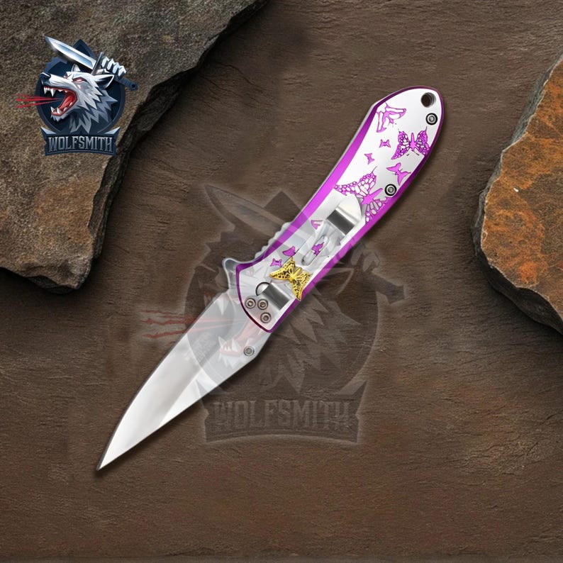Pu&ograve; includere: Un coltello pieghevole con una lama argentata e un manico con un design viola e bianco con accenti a farfalla. Il coltello ha una clip da tasca e un abbellimento a forma di stella color oro. Il logo del marchio "WOLFSMIITH" &egrave; visibile.