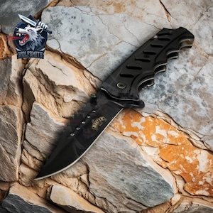 May include: Black folding knife with a serrated blade and a black handle with a textured grip. The knife has a black and gold logo that says "Hunter".