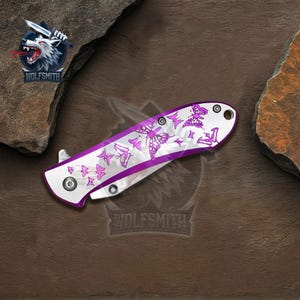 Pu&ograve; includere: Coltello pieghevole con manico bianco e design a farfalle viola. Il coltello ha una lama argentata e un bordo viola. Il logo Wolfsmith &egrave; visibile in alto a sinistra.