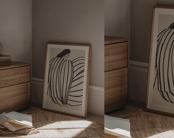 May include: A framed art print featuring a minimalist illustration of a seated figure with striped clothing. The artwork is in black and white, set against a neutral background, and framed in a light wood. The print leans against a wooden cabinet.