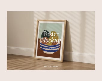 May include: A framed poster mockup featuring an abstract still life of fruit in a bowl. The artwork includes a bowl with blue and white stripes, and the text "Poster Mockup" is visible. The poster is leaning against a wall.
