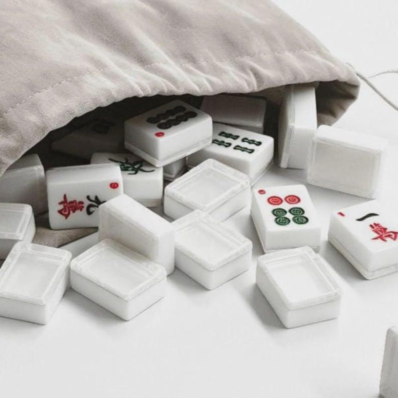 May include: A collection of white Mahjong tiles with black, red, and green markings spilling from a beige drawstring bag. The tiles are rectangular and feature various symbols and characters. The game pieces are scattered on a white surface.