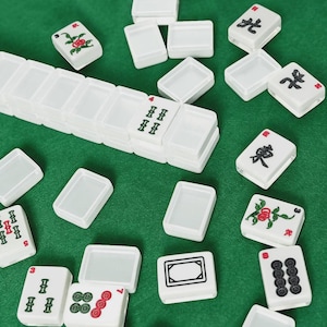 May include: White Mahjong tiles with various symbols and designs, including flowers, numbers, and Chinese characters, are scattered on a green surface. The tiles are rectangular and have a smooth, glossy finish. Some tiles are stacked.