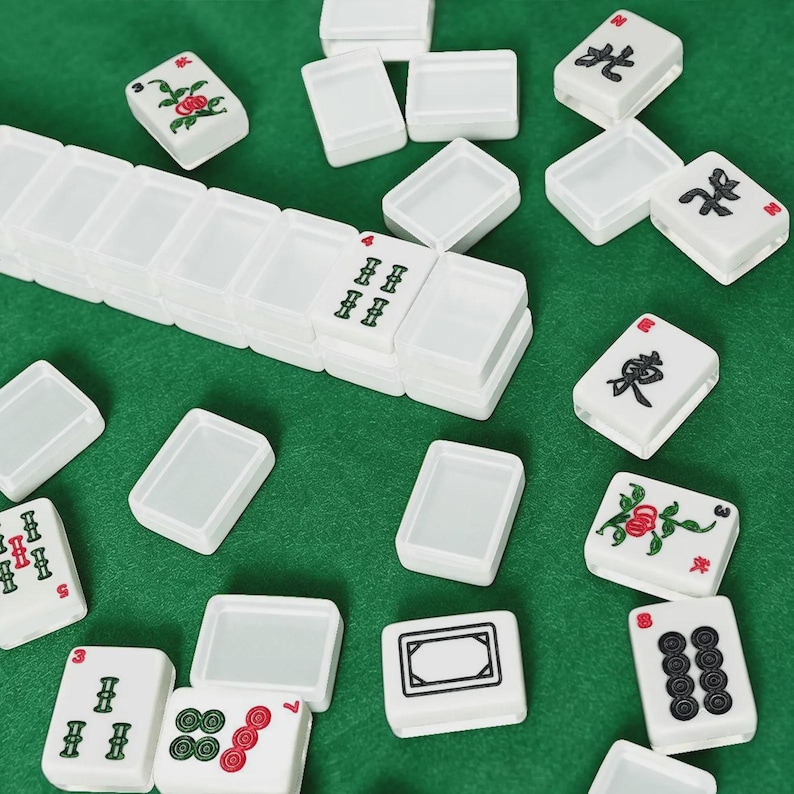 May include: White Mahjong tiles with various symbols and designs, including flowers, numbers, and Chinese characters, are scattered on a green surface. The tiles are rectangular and have a smooth, glossy finish. Some tiles are stacked.